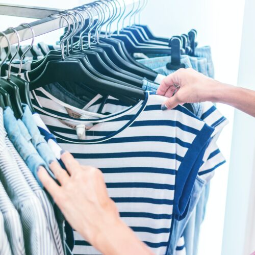 Eine Frau schaut in ihren Kleiderständer nach einem passenden Outfit. Man erkennt ein blau, weiß gestreiftes T-Shirt. Die Dame hat dunkelblauen Nagellack.