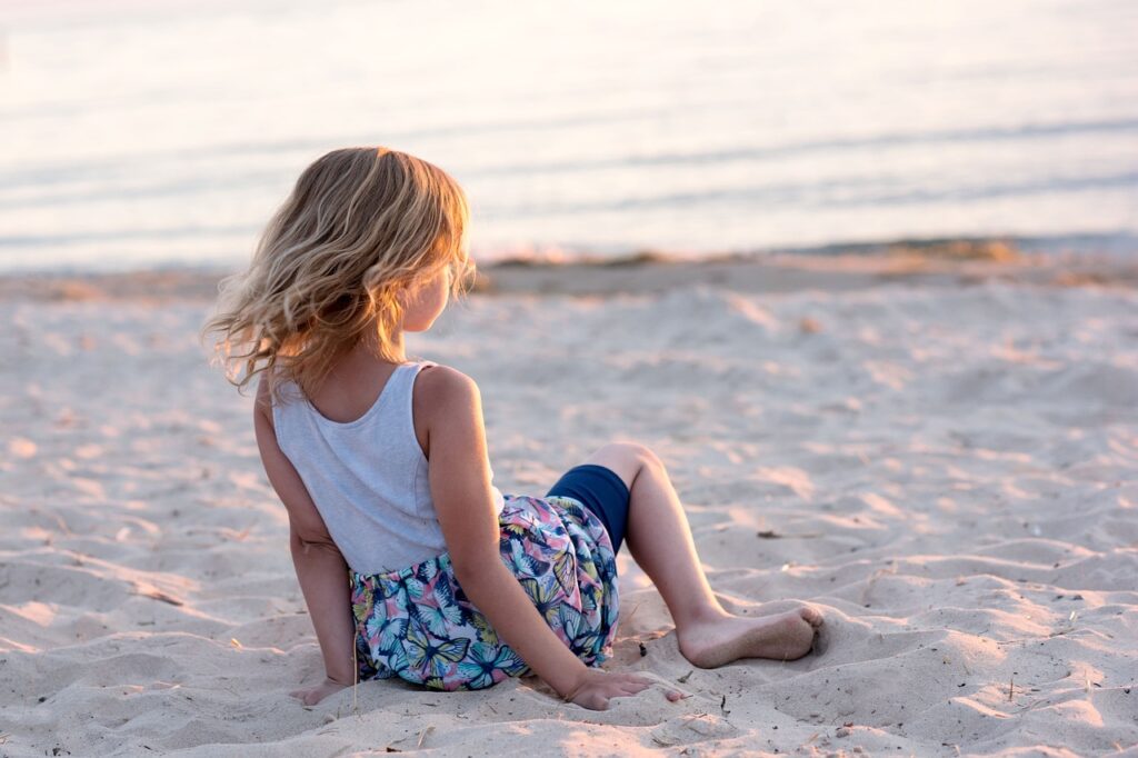 Ein Kind mit blonden langen Haaren sitzt am Sandstrand und schaut in Richtung Sonnenuntergang.