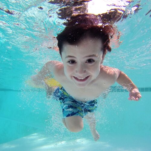 Ein Foto von einem kleinen Kind in einem Pool Unterwasser, was direkt in die Kamera schaut und lächelt.