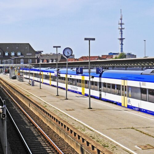 Der HBF auf Sylt. links der Autozug, auf dem ein auto auffährt, rechts ein leers Bahngleis und daneben ein Bahngleis mit einem stehenden Zug. Es ist blauer Himmel, die Bahnuhr zeigt 16:30Uhr an und es ist sonnenschein.