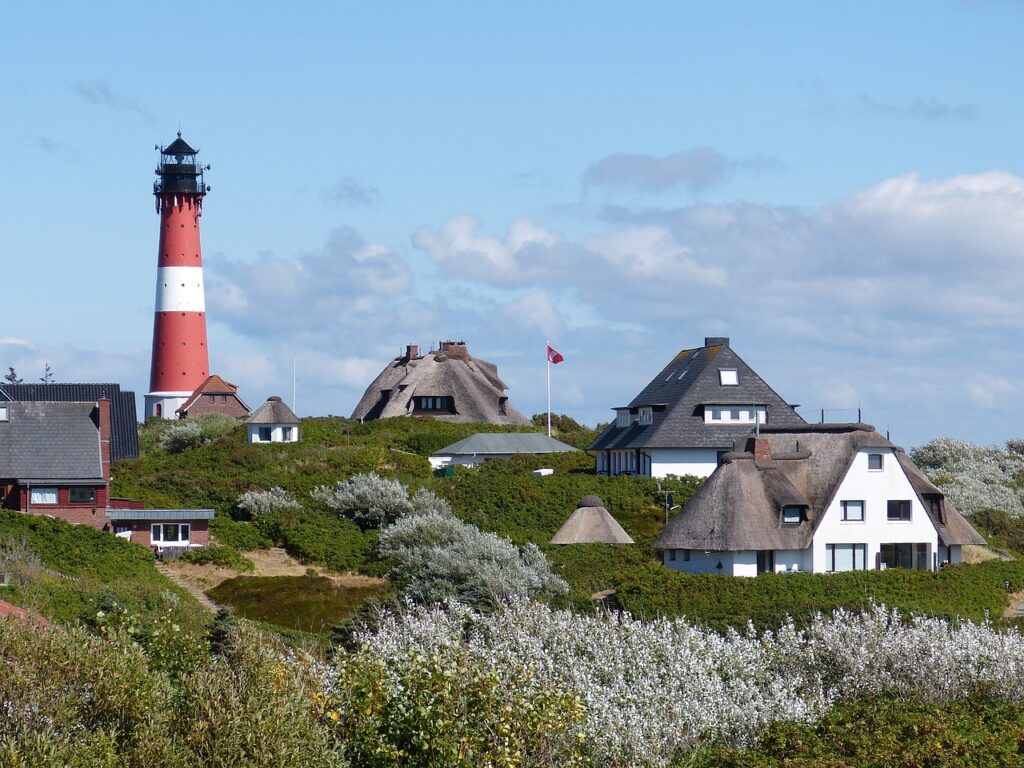 Reetdachhäuser und Leuchtturm in Hörnum auf Sylt. Blauer Himmel im Hintergrund und grüne Wiesen vor den Häusern.