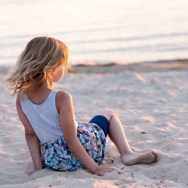 Ein Kind mit blonden langen Haaren sitzt am Sandstrand und schaut in Richtung Sonnenuntergang.