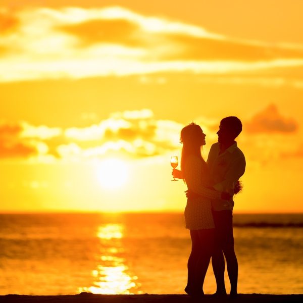 Urlaub zu zweit auf Sylt. Ein Pärchen im Sonnenuntergang am Meer. Die beiden haben ein Weinglas in der Hand und sind nur als Silouhette erkennbar.